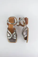 Sandalias de tacón bajo geométrico de madera, confeccionadas en cuero metalizado plateado y detalles de malla transparente, con diseño de tiras orgánicas en la capellada y tira ajustable al tobillo con hebilla metálica.