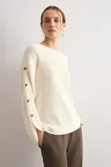 Sweater de punto color blanco natural, con cuello redondo y mangas largas adornadas con botones dorados a lo largo de las costuras exteriores.