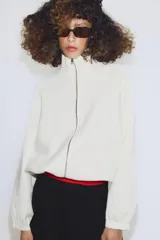 Chaqueta de punto color crudo con cuello alto, cierre frontal de cremallera metálica, mangas largas con puños elásticos y bajo ajustable con cordones.