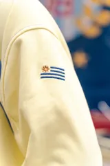 Buzo amarillo de corte holgado con mangas raglán en color azul, detalle de costuras acolchadas en los hombros y bordado de sol en el centro del pecho. Presenta puños y cuello acanalados con rayas azules.