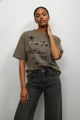 Remera de algodón color gris con efecto lavado, cuello redondo y mangas cortas. Presenta un estampado frontal con nombres de ciudades y detalles de apliques brillantes lineales.