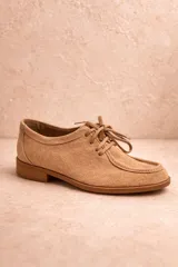 Zapato tipo derby acordonado confeccionado en cuero nobuk color tostado, con suela plana y costuras visibles en la puntera.