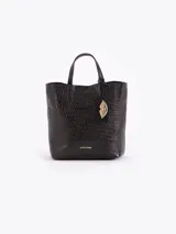 Tote bag Jazmin Chebar de cuero vacuno negro con textura grabada. Presenta un diseño compacto con doble asa superior y un tag colgante con un aplique metálico en forma de labios.