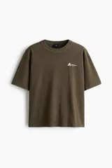 Remera de algodón verde militar con efecto lavado, cuello redondo de canalé, mangas cortas con hombros caídos y pequeño estampado tipográfico en el pecho.
