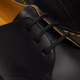 Zapato de cuero negro con cordones, diseño clásico de tres ojales, suela de goma con cámara de aire y costuras amarillas distintivas en el borde.