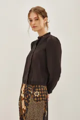 Camisa blanca de algodón con cuello alto, cierre frontal de botones ocultos y detalles de bordado floral en el pecho. Presenta mangas largas con puños elásticos y volados decorativos en la parte frontal.