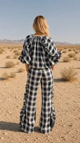 Pantalón cuadrillé de tiro medio con estampa a cuadros en tonos beige, gris y azul. Presenta diseño de perneras anchas, bolsillos laterales y traseros, y detalles de volados con costuras decorativas en zig-zag a lo largo de las piernas.