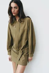 Vestido corto verde militar de cuello solapa y manga larga, con bolsillos de plastrón en el delantero y cierre frontal con cremallera oculta por sopata. Cuenta con un cinturón incorporado del mismo tejido con hebilla metálica y aberturas laterales en el bajo.