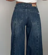 Pantalón jean wide leg de denim azul con efecto lavado, tiro medio-bajo, bolsillos traseros grandes aplicados y costuras verticales decorativas a lo largo de las piernas.