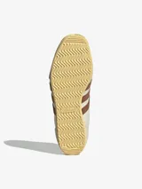 Championes Adidas modelo Japan, con diseño retro de perfil bajo. Presentan un cuerpo de cuero color crema con perforaciones en la puntera, icónicas tres tiras laterales en color marrón y refuerzos de gamuza gris en la puntera y el talón. Incluye suela de goma color amarillento y detalles dorados en el lateral y la lengüeta.