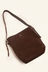 Cartera tipo balde color tostado, confeccionada en gamuza, con correa ajustable decorada con tachas metálicas plateadas.