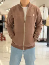 Campera de felpa color marrón, de corte regular, con cierre frontal completo, bolsillos tipo canguro y cuello alto acanalado. Cuenta con un pequeño bordado en el pecho.