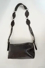 Cartera de hombro color marrón oscuro, confeccionada en cuero sintético con acabado brillante. Presenta una correa larga ajustable con detalles de piezas circulares decorativas y remaches metálicos.