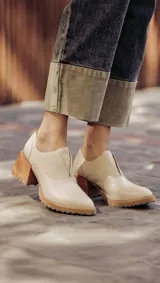 Zapato de cuero color beige, con taco de madera y suela dentada. Presenta un diseño slip-on con detalles de textura en relieve en los laterales y el empeine.