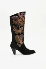 Bota de caña alta confeccionada en gamuza negra, con un diseño artesanal de parches en cueros texturizados y metalizados con motivos florales. Presenta un taco cónico y punta almendrada.