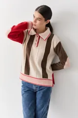 Buzo de punto con cuello polo y cierre frontal de tres botones. Presenta un diseño a rayas verticales en tonos beige y marrón, con mangas en bloques de color rojo y detalles en rosa en los puños y el ruedo.
