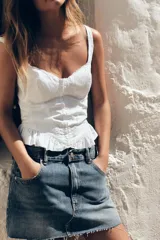 Top blanco estilo corsé con escote corazón, tirantes finos y bordados a tono. Presenta un bajo con volantes y cierre frontal con ganchos metálicos. La espalda es de tejido elástico tipo nido de abeja.