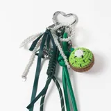 Llavero decorativo con mosquetón en forma de corazón, compuesto por una cadena de perlas, cintas de tela verde, cordones técnicos y un dije tejido a crochet con forma de kiwi.