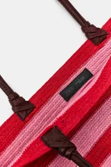 Bolso tipo cesto de yute con diseño de rayas horizontales en tonos rojo y rosado, asas de mano de cuero en color marrón oscuro y base redondeada.