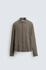 Camisa de hombre de corte relaxed fit, color marrón oscuro, con cuello solapa, manga larga terminada en puño con botón y cierre frontal de botonadura. El tejido presenta una textura acolchada o gofrada con patrón de pequeños cuadros.