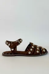 Sandalias planas de gamuza marrón, con tiras cruzadas en el empeine y tira en el tobillo con hebilla ajustable. Están decoradas con tachas metálicas plateadas semiesféricas a lo largo de las tiras y en todo el contorno de la suela.