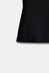 Chaleco negro sin mangas, de cuello redondo y corte entallado con peplum sutil. Presenta pinzas verticales en el frente y falsos bolsillos de vivo a la altura de la cadera.