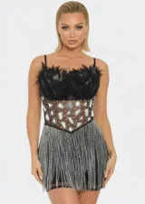Vestido corto de fiesta con escote recto y tirantes finos. El diseño presenta un cuerpo superior decorado con plumas negras y una sección central transparente con apliques de espejos geométricos. La falda está compuesta por una capa de flecos de strass plateados que aportan movimiento y brillo.