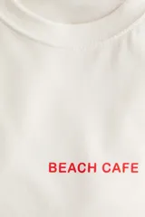 Remera de algodón color crema, con cuello redondo y mangas cortas. Presenta un estampado central en rojo con tipografía cursiva que dice "Papaya", y texto adicional que incluye "Beach Cafe", "Half Moon Bay", "Est 1985" y "Antigua", junto a un pequeño gráfico de una papaya.