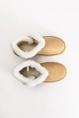 Bota tipo pantubota color beige, confeccionada en material gamuzado con interior y borde superior de piel sintética blanca. Presenta un detalle de botón lateral y suela de goma antideslizante.