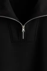 Buzo negro de tejido pesado con interior cepillado suave, cuello alto y cierre frontal parcial plateado. Presenta hombros caídos y puños y bajo en punto acanalado.