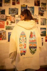 Hoodie color natural con capucha, de corte oversize, que presenta un diseño bordado en la espalda con dos tablas de surf decoradas con motivos playeros y florales, junto con el logo de la marca bordado en la parte inferior.