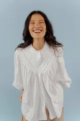 Camisa de algodón color beige con cuello mao, cierre frontal con botones y mangas abullonadas con frunces en los hombros. Presenta costuras verticales decorativas en el frente.