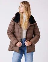 Campera puffer color marrón con capitoneado horizontal. Tiene cuello alto y capucha ajustable con cordones, con el interior del cuello en textura aterciopelada negra. Cierre frontal completo y bolsillos laterales. El corte es ligeramente más largo en la parte posterior con aberturas laterales redondeadas.