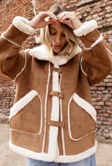 Campera estilo gamulán color tostado con interior y bordes de corderito sintético color crema. Presenta capucha amplia, cierre frontal con trabas y broches metálicos, y bolsillos delanteros grandes. Posee un calce relajado y diseño de textura suave.
