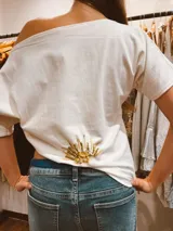 Remera de algodón color blanco con escote asimétrico de hombro descubierto. Presenta un diseño pintado a mano con un sol en la parte inferior y alas en la parte superior.