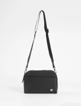 Bandolera de diseño rectangular con doble compartimento y cierre superior. Cuenta con una correa larga ajustable y un bolsillo frontal abierto. Presenta un pequeño aplique metálico con logo en la esquina inferior.