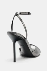 Sandalia plateada de tacón fino, con tira ancha en la parte delantera y pulsera al tobillo con cierre de hebilla metálica. Zapato con acabado en punta y efecto metalizado.