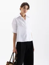 Camisa de popelina blanca con bordados de pequeñas bocas que forman un patrón de rayas. Presenta un diseño de corte holgado, mangas cortas con volumen, cuello clásico con broche metálico y cartera oculta con botones al tono.