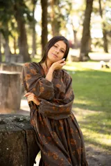 Vestido largo de lino con escote en V, mangas largas abullonadas y falda con volado en el ruedo. Presenta un estampado floral en tonos tierra sobre base marrón.