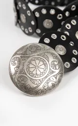 Cinto de cuero negro compuesto por piezas circulares unidas, decoradas con ojales metálicos plateados y apliques circulares grabados. Presenta una hebilla central grande de metal plateado con diseño ornamental grabado.