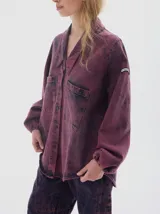 Camisa oversized de denim con lavado acid wash en tonos morados y azul oscuro. Presenta cuello camisero, escote en V, dos bolsillos frontales con tapa y botones oscuros. Las mangas son largas tipo balloon con puño ajustado. El bajo es irregular.