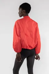 Campera bomber color rojo anaranjado de nailon repelente al agua, corte regular, cierre frontal con cremallera visible, tirador con logo bordado, bolsillos de ribete verticales, hombro caído, bolsillo utilitario con cremallera en la manga, cuello de chaqueta bomber acanalado y puños y bajo elásticos fruncidos.