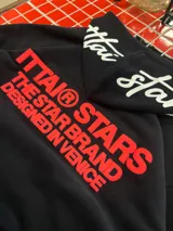 Hoodie negro de corte oversize con capucha, confeccionado en algodón. Presenta un estampado gráfico en color rojo en la espalda con la leyenda 'ITTAI STARS THE STAR BRAND DESIGNED IN VENICE' y un detalle bordado en blanco en la parte posterior de la capucha.