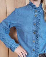 Camisa de denim con volados verticales a lo largo de la botonadura frontal y cuello alto. Presenta mangas largas con puños elásticos y detalle de volados en los mismos.