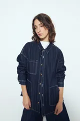 Sobrecamisa de jean color azul oscuro con estampado de rayas verticales blancas. Presenta un corte relajado, cuello clásico, cierre frontal con botones metálicos, mangas largas y dos bolsillos frontales de parche.