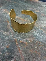 Pulsera rígida tipo brazalete de diseño ancho y textura irregular con acabado dorado brillante.