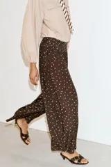Pantalón de tiro alto con pernera ancha y diseño fluido. Presenta un estampado de lunares blancos sobre fondo marrón y detalle de frunces en la cintura.