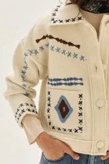 Campera de lana merino hervida en color crudo, con diseño de cuello camisero, cierre frontal de botones y bolsillos frontales. Presenta bordados artesanales con motivos geométricos en tonos marrón y azul en el frente, espalda y mangas.