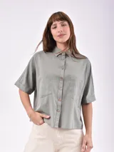 Camisa de manga corta, color verde oliva, con cuello camisero y botones de madera. Presenta dos bolsillos frontales con tapa y un corte holgado.