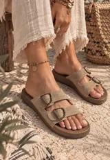 Sandalias planas de mujer con doble tira en tono mostaza de acabado gamuzado, sujetas por dos hebillas metálicas de estilo rústico y base marrón.